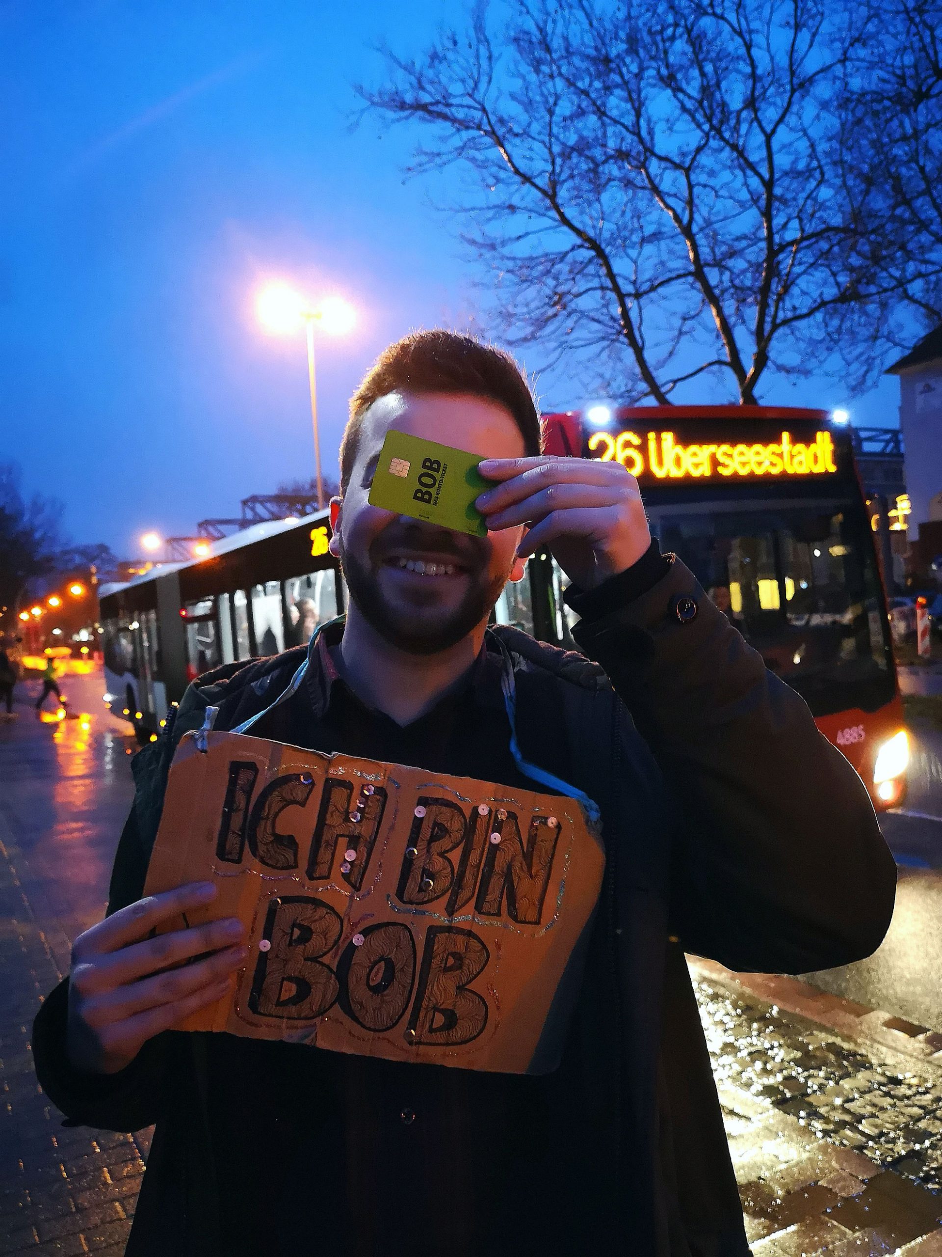 Ein junger Mann hält sich mit seiner linken Hand eine grüne Plastik-Karte im EC-Karten-Format vor die Augen: Das BOB-Konto-Ticket der BSAG. Mit der anderen Hand hält er sich ein Pappschild vor die Brust mit der Aufschrift: "Ich bin BOB". Im Hintergrund fährt ein BSAG-Gelenkbus Linie 26 Überseestadt.