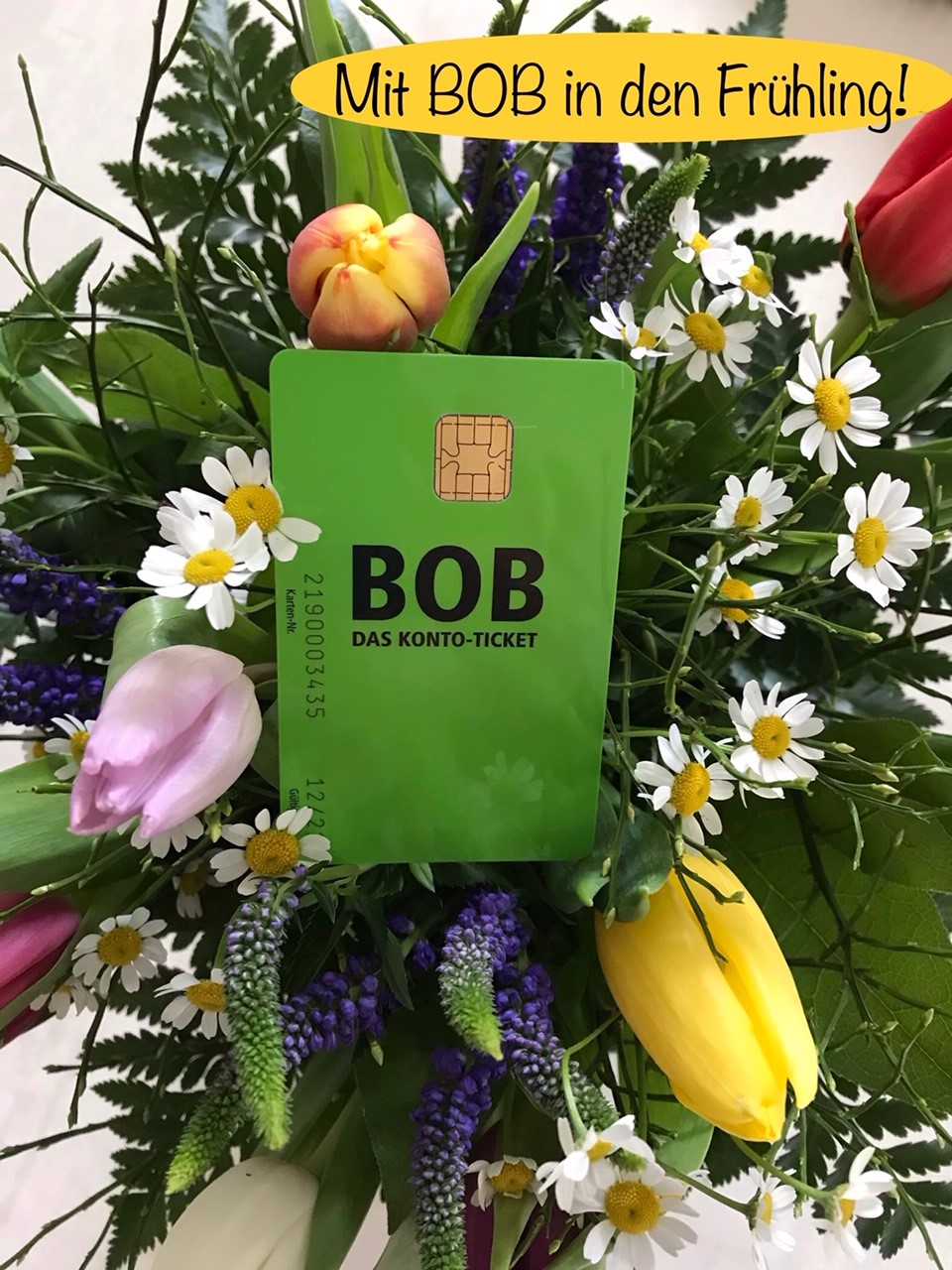 Eine grüne Plastik-Karte im EC-Karten-Format liegt in einem von oben fotografierten Frühlings-Blumenstrauss: Das BOB-Konto-Ticket der BSAG. Rechts oben im Bild steht in schwarzer Schrift auf gelbem ovalen Hintergrund "Mit BOB in den Frühling!"