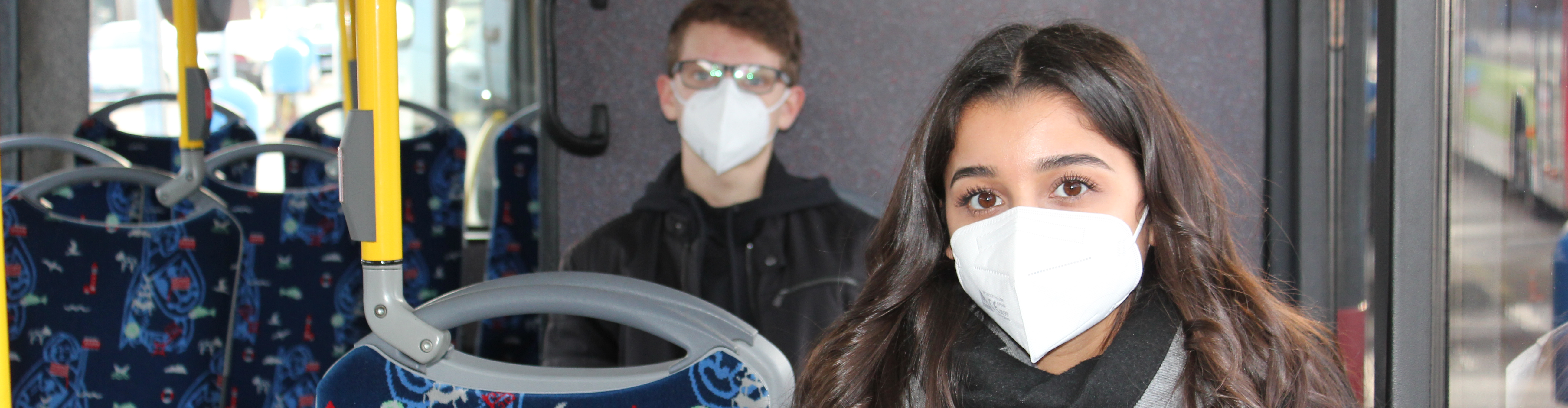 Ein junger Mann und eine junge Frau sitzen im Bus. Beide tragen eine FFP2-Maske.